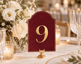Numéros de table en velours, détails subtils en fil doré, découpés à l'emporte-pièce, supports dorés inclus, décoration de table élégante pour un mariage.