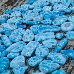 Peut inclure: Une collection de pierres précieuses Larimar polies, bleu clair et blanc, de formes et de tailles variées, notamment ovales, en goutte d'eau et rectangulaires. Chaque pierre présente un motif marbré unique. Le texte BULKDRUZYGEMS est visible.