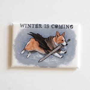 May include: Rectangular magnet featuring a corgi dog illustration with a sword and wings, running across a blue and gray background. The text "WINTER IS COMING" is above the dog. The corgi is brown, white, and black.