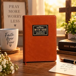 May include: A rust-colored notebook with a black rectangular label on the front cover featuring the gold text "WITH GOD ALL THINGS ARE POSSIBLE". The notebook has a smooth, solid-colored cover and a thin ribbon bookmark extending from the bottom edge.