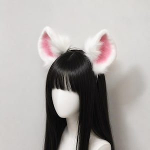 以下が含まれることがあります： ふわふわした白いフェイクファーと柔らかなピンク色の中心部を持つ、2つの大きな動物の耳が付いたヘッドバンド。耳は細いダークカラーのヘッドバンドフレームに取り付けられており、ストレートの黒髪と前髪を持つマネキンの頭部に装着されています。