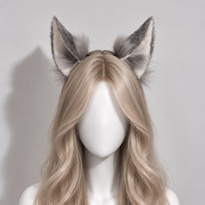 May include: A pair of upright, pointed animal ears made of faux fur in shades of grey and white, attached to a thin black headband. The ears feature a fluffy texture with darker grey tips and lighter white inner sections, positioned on a mannequin head.