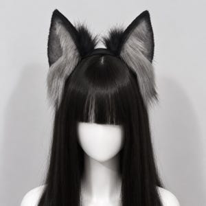 May include: A headband featuring two tall, pointed animal ears covered in long, textured faux fur. The ears are primarily black with grey highlights along the inner edges and base, attached to a simple black band worn on a mannequin head.