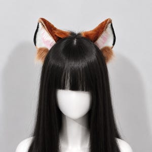 Puede incluir: Un par de orejas de animal de piel sintética en tonos marrón, negro y blanco, con puntas suaves y detalles en rosa claro en el interior. Las orejas están unidas a una diadema, mostradas sobre una cabeza de maniquí con cabello negro largo.