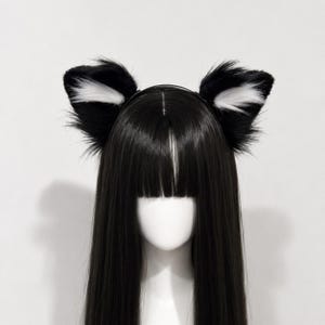 May include: A headband featuring two plush, pointed animal ears made of black faux fur with white fur accents on the inner sections. The headband is displayed on a mannequin head with long, straight black hair that has blunt-cut bangs.