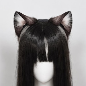 Pode incluir: Um par de orelhas de gato de pele sintética em tons de preto, cinza e branco, presas a uma tiara preta. As orelhas apresentam um padrão de cor degradê com pontas escuras e seções internas mais claras, posicionadas sobre uma peruca preta.