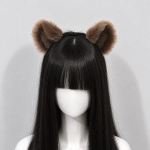 Puede incluir: Una diadema negra con dos orejas de animal redondeadas y erguidas, cubiertas de suave piel sintética marrón. Las orejas están colocadas simétricamente en la parte superior de la banda, mostrando un pelo denso y texturizado que imita la piel natural.