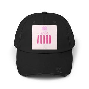May include: A black baseball cap with a distressed brim and a square patch on the front. The patch features a light pink background with a pink brain illustration at the top, the script text Authentically, and the block text AUDHD in pink letters.