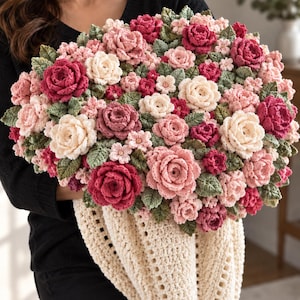 May include: A large, dense bouquet of crocheted flowers in shades of deep magenta, soft pink, and cream, interspersed with textured green crocheted leaves. The floral arrangement is gathered into a cream-colored, open-weave crocheted fabric wrap.