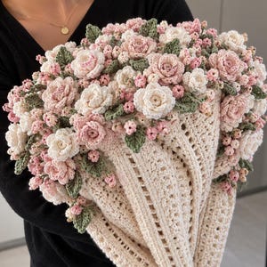 May include: A cream-colored crocheted wrap or shawl featuring an elaborate floral arrangement of pink and white crocheted roses, small buds, and green leaves attached to the upper edge, creating a dense, textured, and layered botanical design.