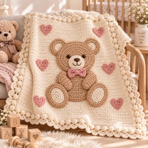 Puede incluir: Una manta de ganchillo color crema que presenta un oso de peluche marrón grande centrado con una pajarita rosa. La manta está decorada con varios corazones pequeños de ganchillo rosa y un borde texturizado de puntos de bodoque.
