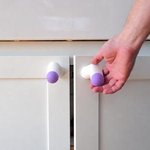 May include: Two white cabinet knobs with rounded purple tips attached to white shaker-style cabinet doors. The knobs feature a cylindrical white base that extends outward, ending in a smooth, spherical purple cap, providing a grip for opening.