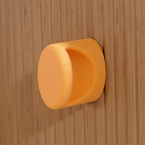 May include: A light orange, circular, textured cabinet knob attached to a light brown wooden drawer front. The drawer front features a vertical fluted or ribbed pattern with a visible wood grain texture throughout the surface.