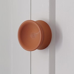 May include: A terracotta-colored circular cabinet knob with a concave center and a smooth, matte finish. The knob is mounted on a white paneled surface, featuring a distinct rounded rim and a cylindrical base that extends from the center.