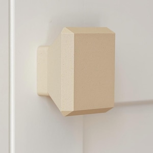 May include: A cream-colored cabinet knob with a rectangular, faceted front face and a tapered base. The hardware features a matte, slightly textured finish and a geometric design with beveled edges, mounted securely to a flat surface.
