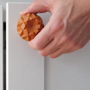 May include: A round, terracotta-colored cabinet knob featuring a complex, multi-faceted geometric pattern with triangular surfaces. The knob is attached to a smooth, light gray cabinet door, showcasing a textured, matte finish on its surface.