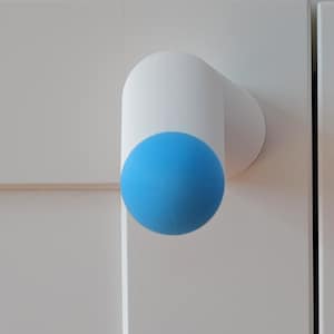 May include: A white cylindrical cabinet knob featuring a bright blue spherical end cap. The knob is mounted onto a white panel surface, showcasing a clean, geometric design with a smooth finish and a distinct contrast between the white and blue.