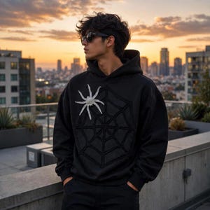 Op de afbeelding: Een zwarte hoodie met een groot, gestikt spinnenwebontwerp op de borst en een opvallende witte spinnenapplicatie aan de rechterbovenkant van het web. Het kledingstuk is voorzien van lange mouwen, een capuchon en een geribbelde zoom.