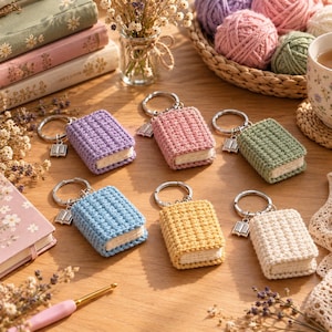 Könnte beinhalten: Sechs gehäkelte Buch-Schlüsselanhänger in Lila, Rosa, Grün, Blau, Gelb und Creme, jeweils mit einem silbernen Metallring und einem kleinen silbernen Buchanhänger. Die Miniaturbücher haben strukturierte Einbände und cremefarbene Seiten.