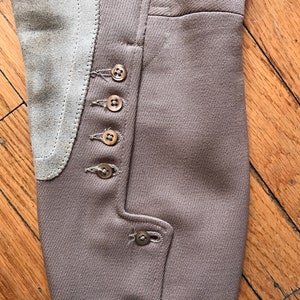 May include: Close-up of the brown fabric leg of a pair of riding breeches with a suede knee patch and multiple brown buttons.