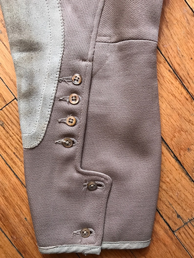 May include: Close-up of the brown fabric leg of a pair of riding breeches with a suede knee patch and multiple brown buttons.