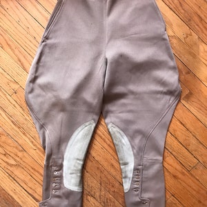 May include: A pair of beige riding breeches with white leather knee patches and button closures at the bottom of the legs.