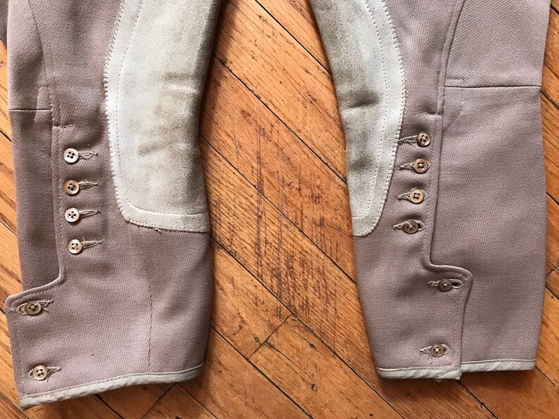 May include: A pair of brown riding breeches with suede knee patches and multiple button closures on the sides. The breeches have a white trim around the bottom.