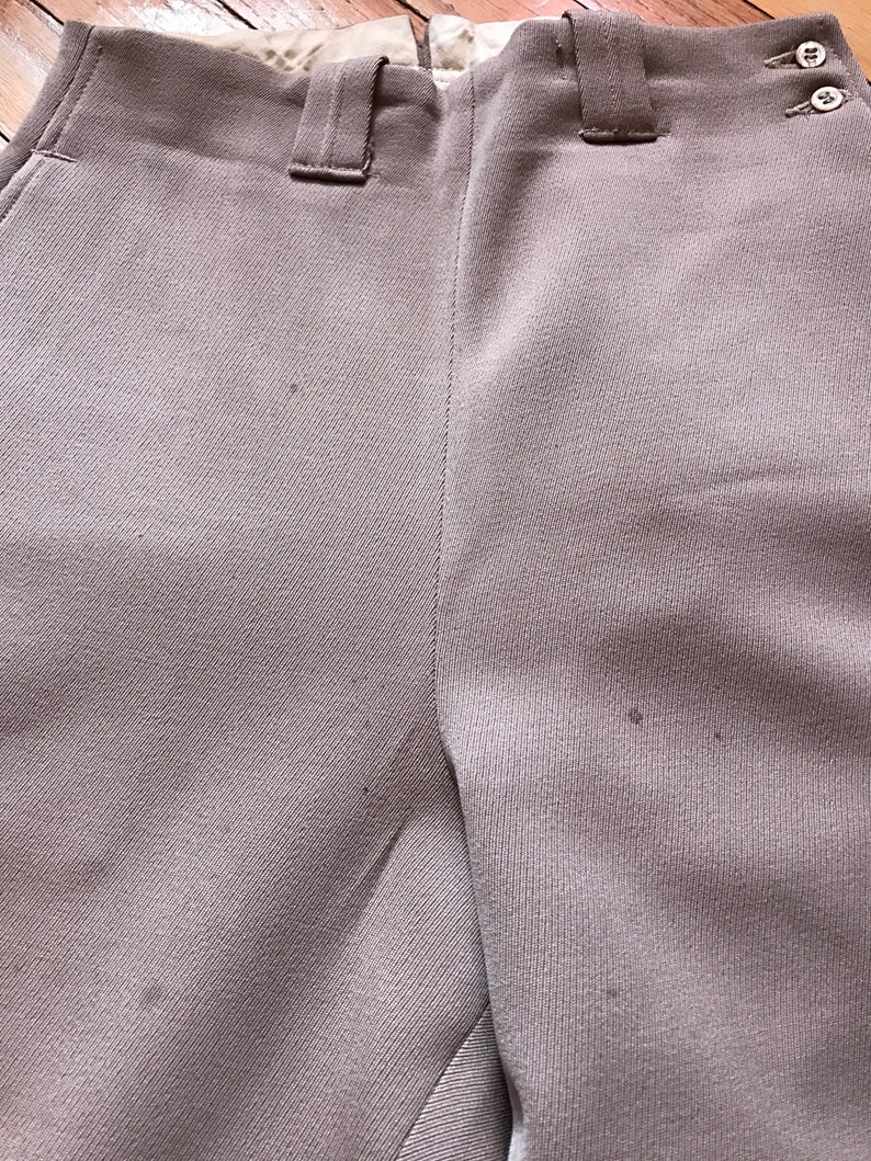 May include: A pair of light brown pants with a button fly and belt loops. The pants are made of a smooth, woven fabric.