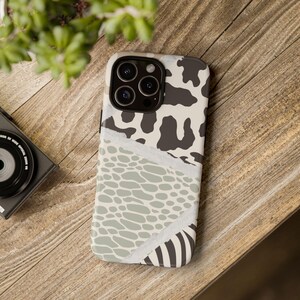 Peut inclure: Une coque de smartphone présentant un motif varié avec un imprimé vache noir et blanc sur le haut, une texture façon peau de serpent vert sauge et crème au centre, et des rayures zébrées noires et blanches en bas, séparées par des lignes blanches.