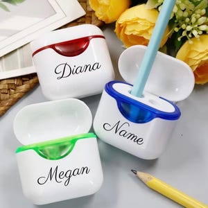 Può includere: Tre temperamatite in plastica bianca a forma di custodie per auricolari, con coperchi colorati in rosso, blu e verde. Ogni temperamatite è personalizzato con testo in corsivo nero che riporta Diana, Name e Megan. Una matita azzurra è inserita.