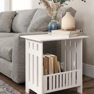 May include: A white side table with a rectangular top and vertical slat side panels. The table features a lower storage compartment containing several books. The top surface holds a blue vase with dried flowers and a ribbed beige diffuser.