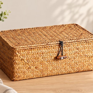 May include: A rectangular storage box crafted from woven light brown seagrass. The box features a flat lid and a secure closure mechanism consisting of a black elastic cord looped around a small, polished wooden toggle button on the front.