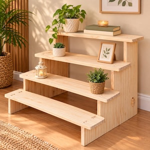 May include: A four-tiered wooden display stand with a light natural finish. The steps hold a potted trailing plant, a small succulent, a white metal lantern, a woven basket planter with a green plant, a picture frame, and two stacked books.