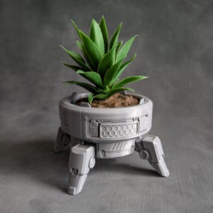 Op de afbeelding: Een groene vetplant in een lichtgrijze, ronde plantenbak met een mechanisch ontwerp. De plantenbak heeft een zeshoekig rasterpatroon aan de zijkant en wordt ondersteund door drie scharnierende, robotachtige poten die naar buiten staan.