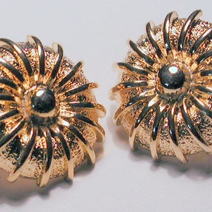 May include: A pair of gold-toned earrings with a sunburst design. The earrings are round and have a textured surface.