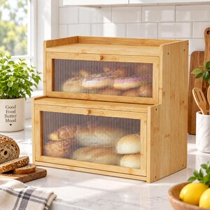 May include: A two-tier bamboo bread box featuring a light wood frame and two drop-down doors with vertical ribbed glass panels. The interior compartments contain various baked goods, including round loaves and rolls, visible through the glass.