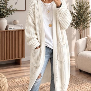 May include: A long, open-front white knit cardigan featuring a chunky ribbed texture, long sleeves with ribbed cuffs, and two large patch pockets on the front. The garment is worn over a plain white t-shirt and light blue distressed denim jeans.