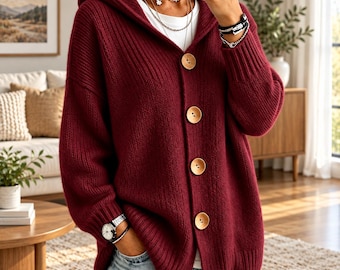 Op de afbeelding: Een diepbordeauxrood vest met capuchon, voorzien van een geribbelde breitextuur en vier grote, ronde, lichtgekleurde houten knopen aan de voorzijde. Het kledingstuk heeft lange mouwen met geribbelde boorden en wordt over een wit shirt gedragen.