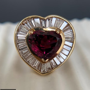 May include: A gold ring featuring a central heart-shaped deep red gemstone surrounded by a halo of tapered baguette-cut clear stones. The setting is heart-shaped, and the piece includes a small logo in the corner with the text RITVIK CREATION.