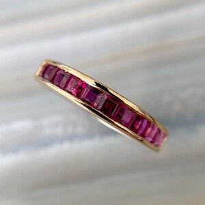 May include: A gold band ring featuring a channel setting of multiple square-cut, deep pink rubies arranged in a continuous row. The metal has a polished finish, and the stones are set flush within the gold frame, creating a sleek, uniform look.