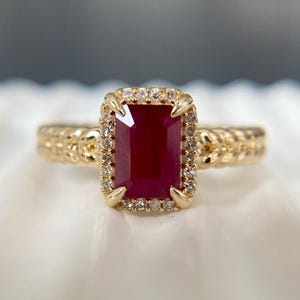 May include: A gold ring featuring a rectangular, deep red ruby center stone surrounded by a halo of small, clear round diamonds. The ring band has a textured, braided metal design. A small logo with the text Soulmates Jewels is in the corner.