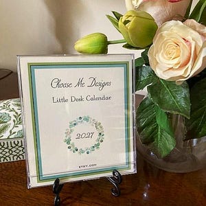 May include: A square desk calendar in a clear plastic case on a black metal stand. The white card features the text Choose Me Designs, Little Desk Calendar, 2027, and ETSY.COM, with a green and blue border and a circular floral wreath graphic.