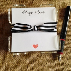 May include: A clear acrylic notepad holder with a black and white striped ribbon bow. The notepad has the name "Mary Anna" printed at the top and a red heart in the center.