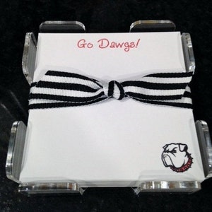 May include: A white notepad with a black and white striped ribbon tied around it. The notepad has the text "Go Dawgs!" printed on the top and a bulldog graphic on the bottom. The notepad is in a clear acrylic holder.