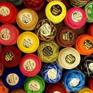 Puede incluir: Una colección de ovillos de hilo de algodón de la marca Butterfly en varios colores sólidos y matizados. Cada ovillo presenta una etiqueta de papel circular con el texto Butterfly, 10g, y Perlé Cotton o Gold, junto a un icono de mariposa.