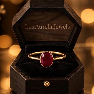 Könnte beinhalten: Ein goldener Ring mit einem glatten, ovalen, tiefroten Edelstein, präsentiert in einer dunkelbraunen, sechseckigen Holzschatulle. Der Innendeckel der Schatulle zeigt den Text LuxAureliaJewels in heller Schrift, der Ring liegt auf schwarzem Samt.