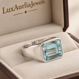Könnte beinhalten: Ein silberfarbener Ring mit einem großen, rechteckigen, hellblauen Edelstein in einer polierten Metallschiene. Der Ring befindet sich in einer braunen Schmuckschatulle, auf deren Innendeckel der Text LuxAurliaJewels in Braun aufgedruckt ist.