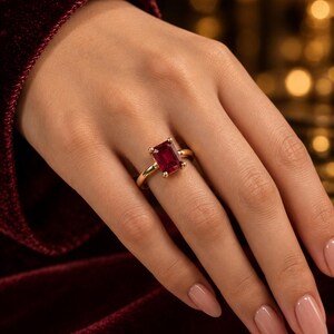 May include: A gold ring featuring a rectangular emerald-cut deep red gemstone set in a four-prong setting. The band is a smooth, polished gold metal. The ring is positioned on a finger, showcasing the vibrant red stone and the gold band.