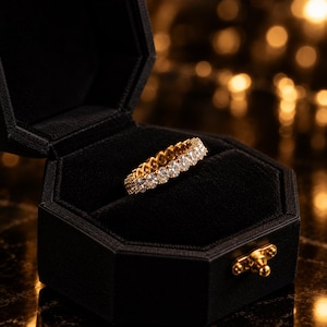 Könnte beinhalten: Ein goldener Eternity-Ring mit einer durchgehenden Reihe runder, klarer, facettierter Steine in einer Metallfassung. Der Ring befindet sich in einer offenen, achteckigen, schwarz samtgefütterten Schmuckschatulle mit einem goldenen Verschluss.