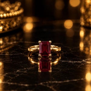 May include: A gold ring featuring a rectangular emerald-cut deep red gemstone set in a four-prong setting. The band is smooth and polished gold, reflecting light on its surface. The gemstone is positioned centrally, showcasing its faceted edges.